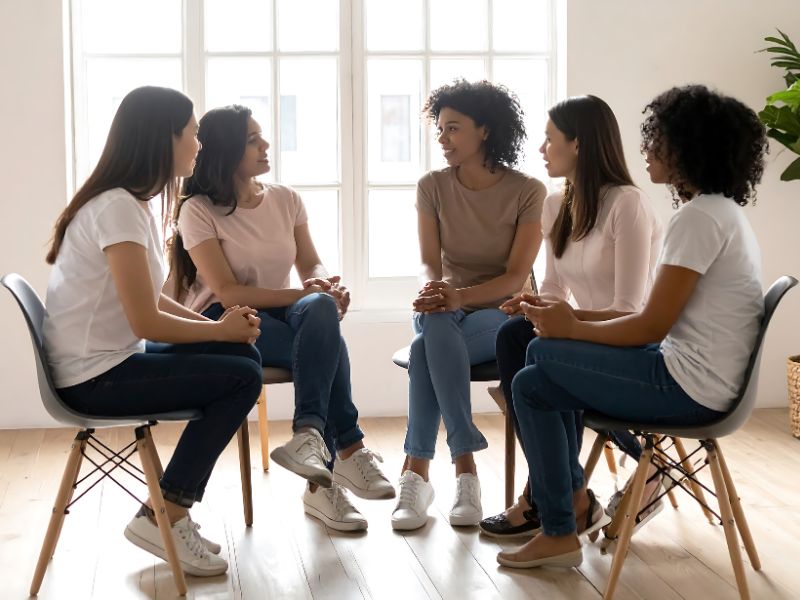 Small group of women seated in a circle in a bright room, talking and listening during a supportive therapy session. Image representing a mental health Partial Hospitalization Program (PHP), offering structured daytime treatment, counseling, and peer support.